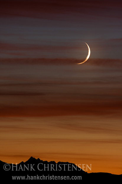 A crescent moon glows in the warm colors of sunset as it sets behind the crest of the Sierra.