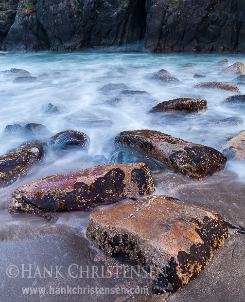 Large flat rocks lead through the rising tide to a dark sea arch in a rock wall, Harris State Beach