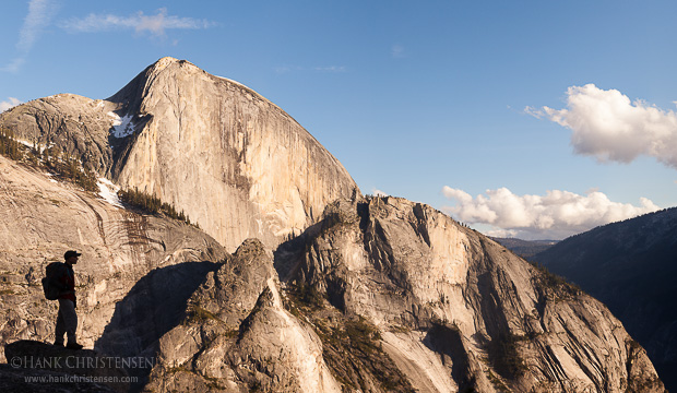 A backpacker stands on an outcropping admiring the view while Half Dome rises high overhead, Yosemite National Park