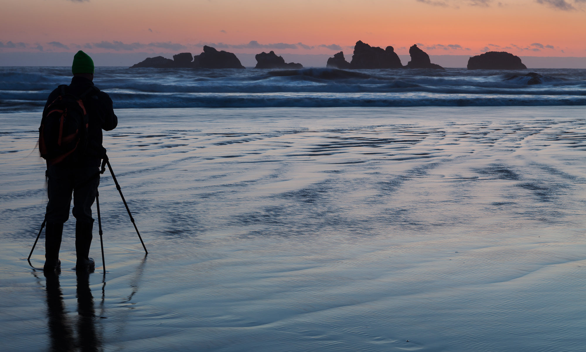 A photographer stands in shallow water filming a fleeting sunset